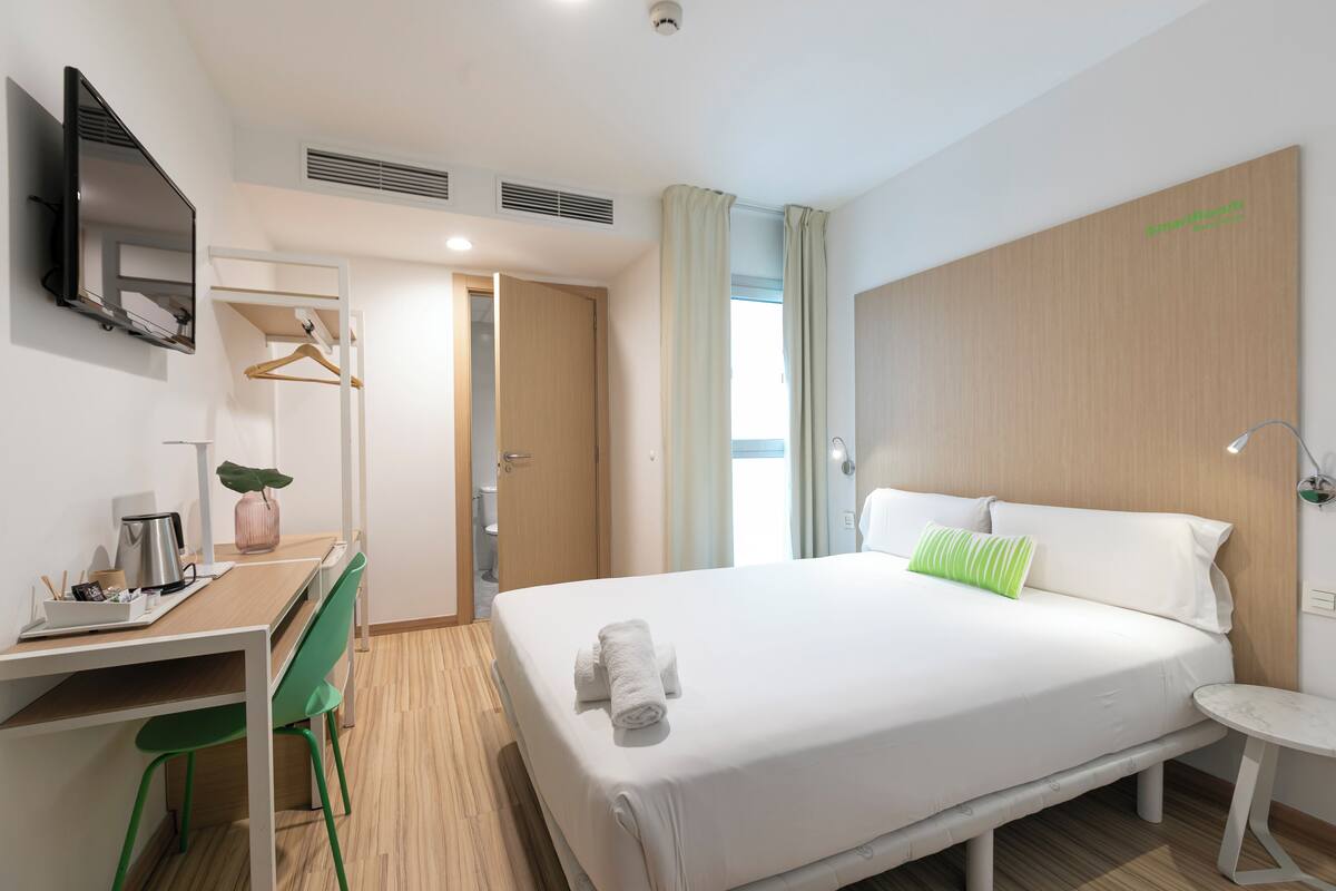 Una camera d'albergo con un letto, una scrivania, una sedia, una televisione e un comodino.