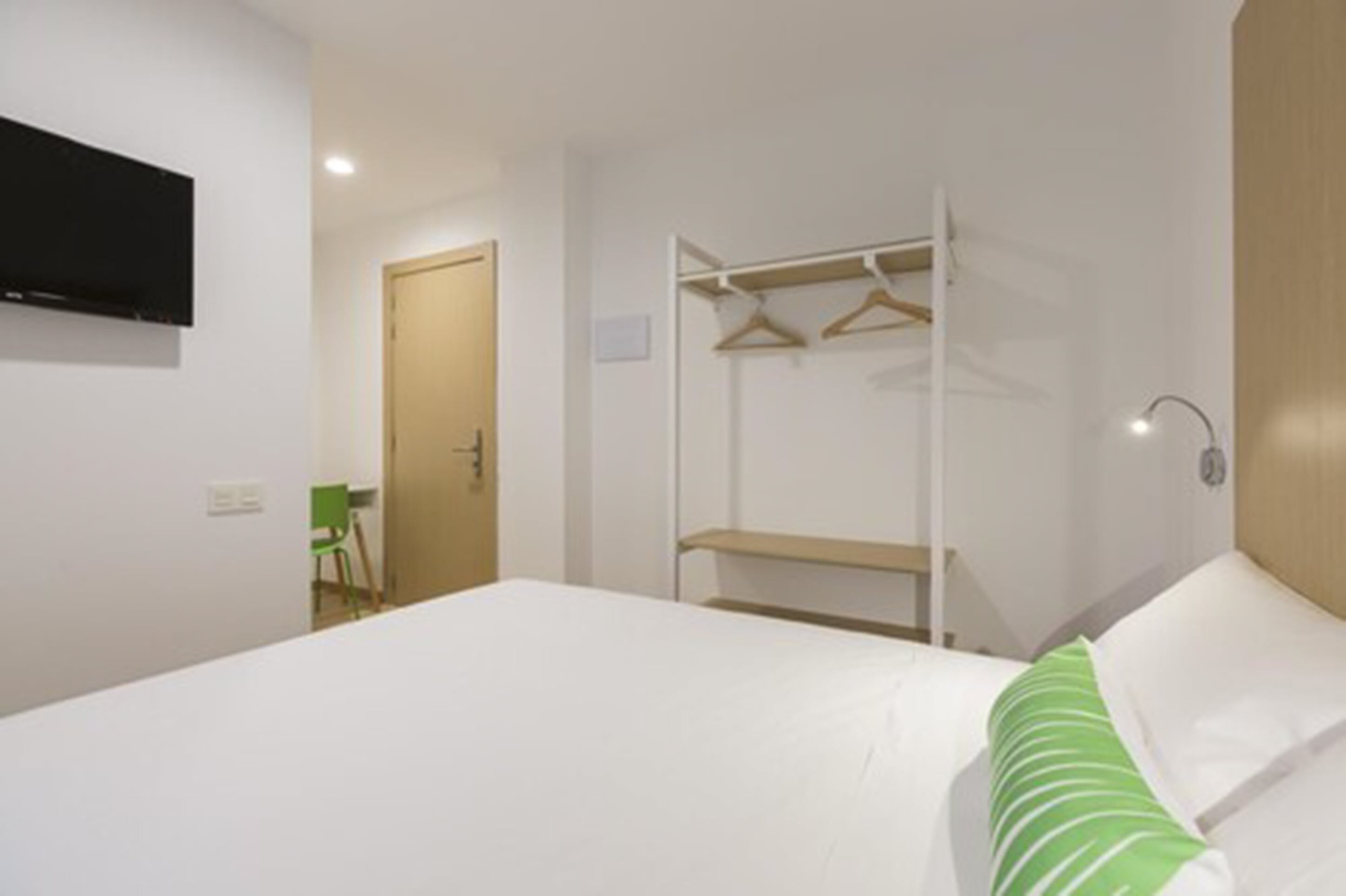 Habitación de hotel con cama, armario con perchas, televisor montado en la pared y una silla verde.