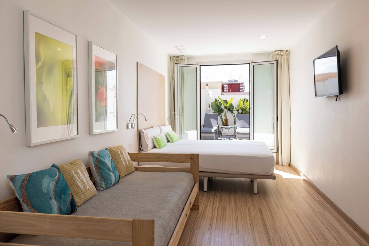 Una camera d'albergo moderna con un letto, un divano, una TV e ampie finestre vista sulla città.