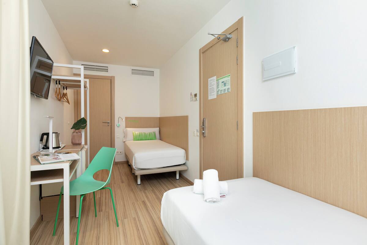 Una camera d'albergo con un letto, una scrivania con una sedia, una televisione a parete e una porta con un cartello.