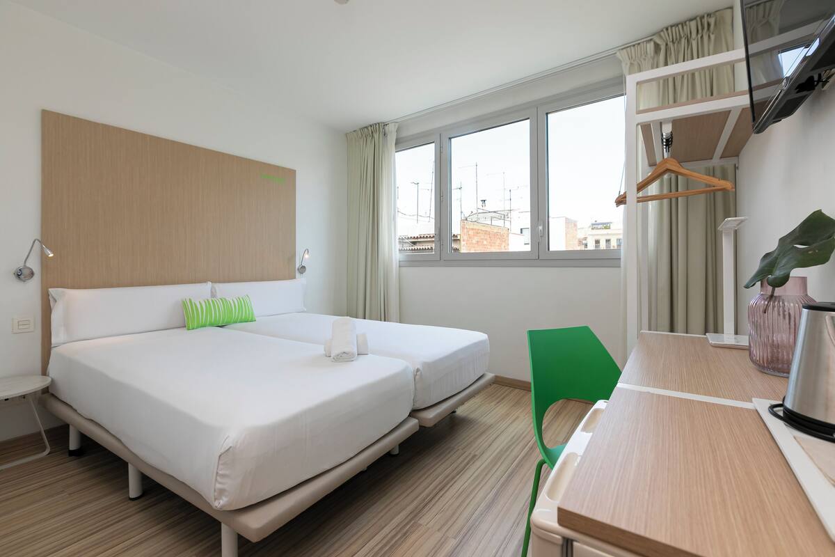 Camera d'albergo con due letti, una scrivania, una sedia, un bollitore e una finestra con tende.