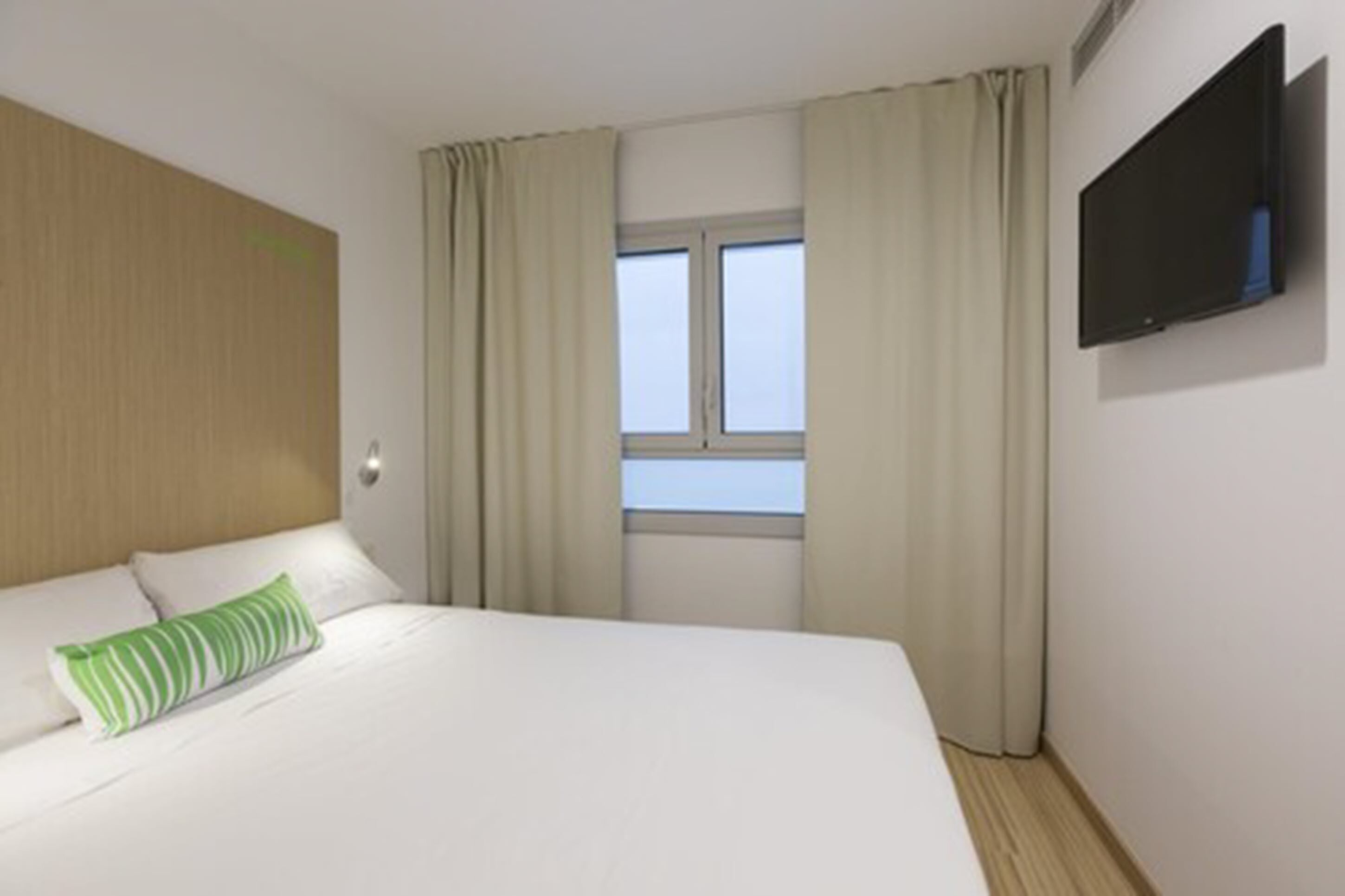 A hotel room with a bed, a television mounted on the wall, and a window with curtains.