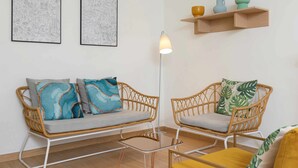Una sala de estar con dos sofás de mimbre, una mesa de centro de vidrio y una lámpara de pie. Hay dos obras de arte enmarcadas en la pared y una estantería con objetos decorativos.