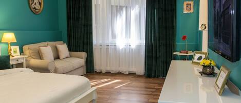 Habitación de hotel con cama, sofá, escritorio con televisor, ventana con cortinas y una mesita de noche con lámpara y marcos de fotos.