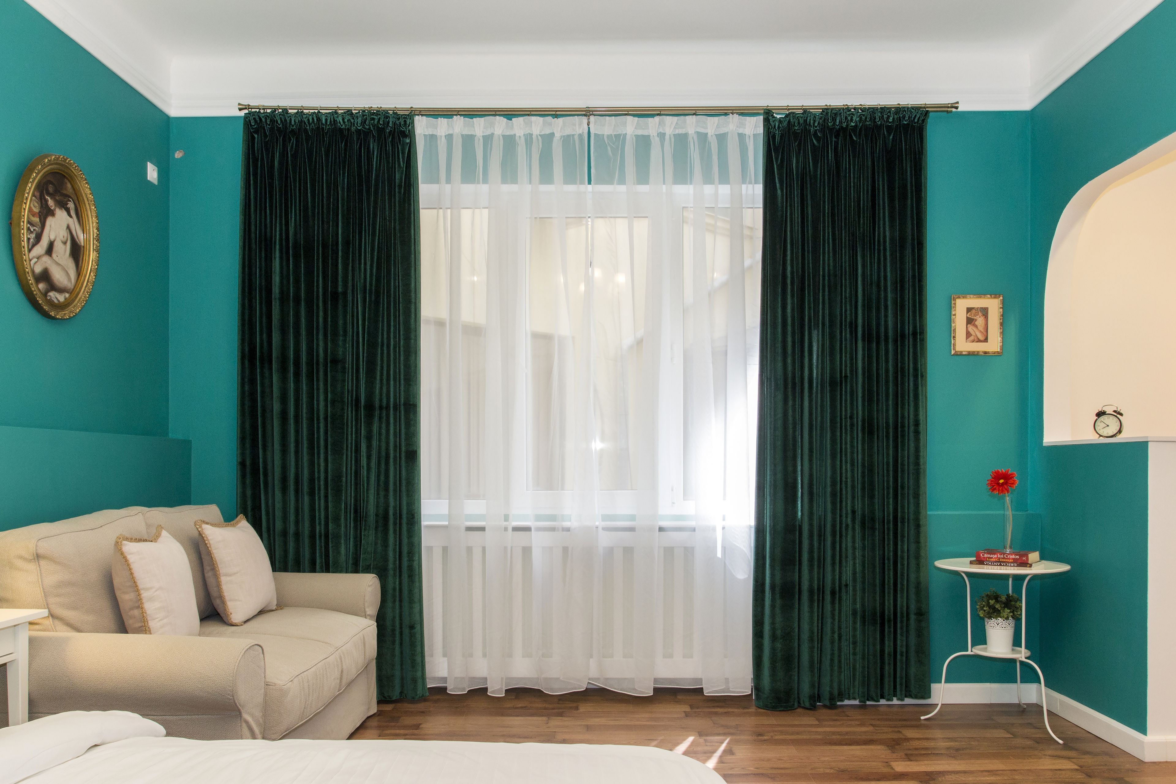Una habitación con paredes verde mar, un sofá color beige, una cama blanca y una ventana con cortinas blancas traslúcidas y cortinas verde oscuro.