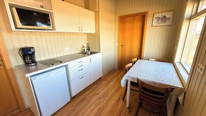 Cocina compacta equipada con microondas, cafetera y un refrigerador pequeño. Hay una mesa de comedor de madera con sillas y una ventana con vista a una casa.