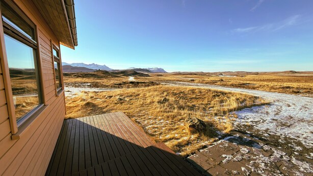 Una terraza de madera que conduce a un ventanal con vista a un paisaje seco y cubierto de hierba, con montañas al fondo.