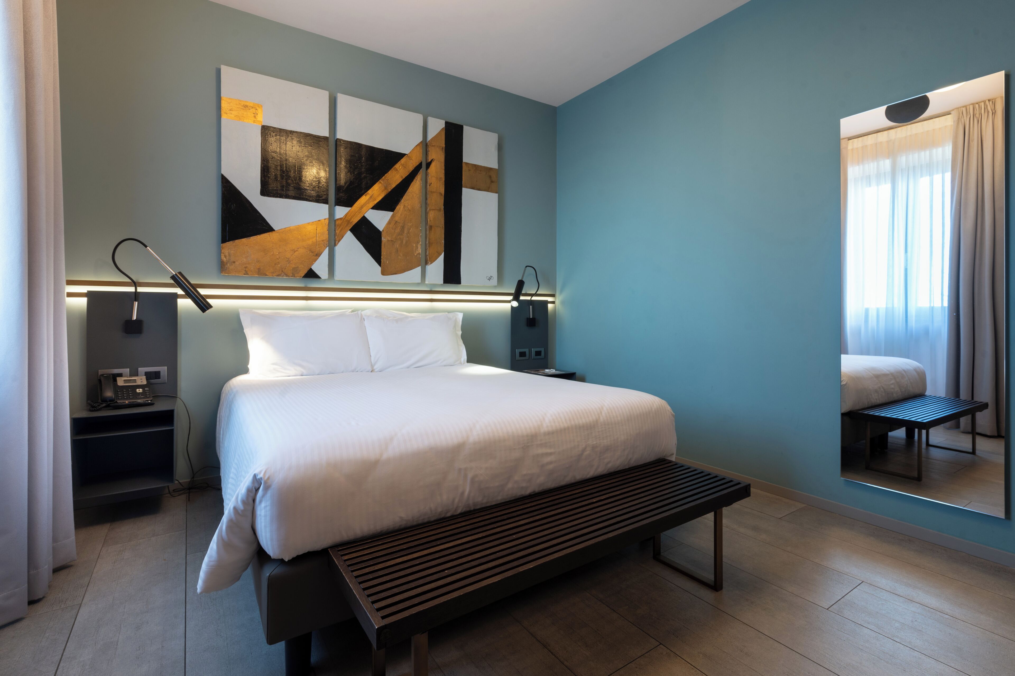 Une chambre d’hôtel moderne dotée d’un grand lit, de tables de chevet, d’un banc, d’un miroir mural et d’œuvres d’art abstraites.