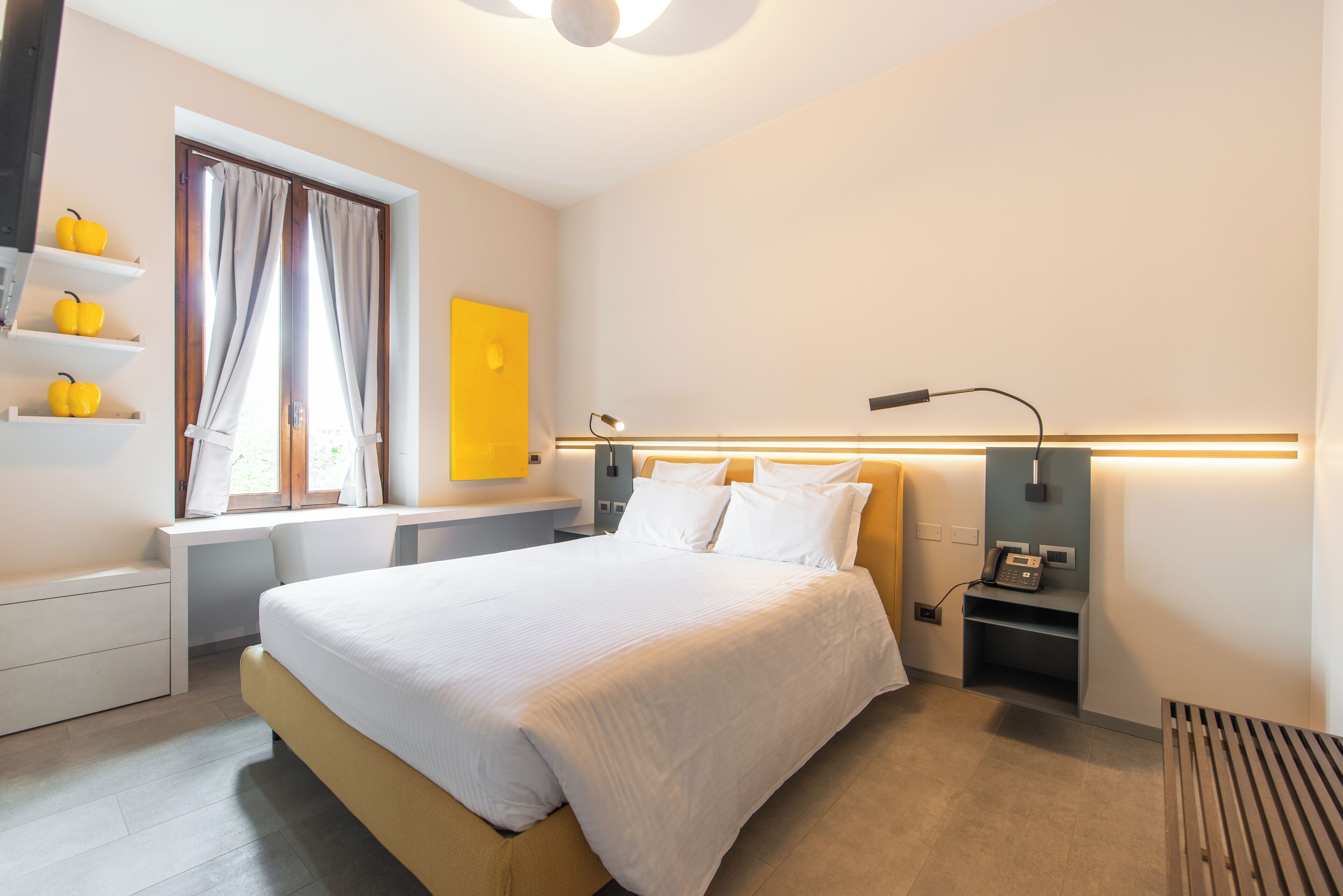 A modern hotel room with a bed, bedside tables, a desk, a lamp, a phone, and a window with curtains.