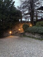 Un sentiero sterrato conduce a una scala in pietra con ringhiera metallica, immersa tra alberi e vegetazione.