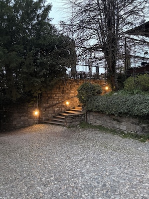 Un sendero de grava conduce a una escalera de piedra con barandilla de metal, rodeado de árboles y vegetación.