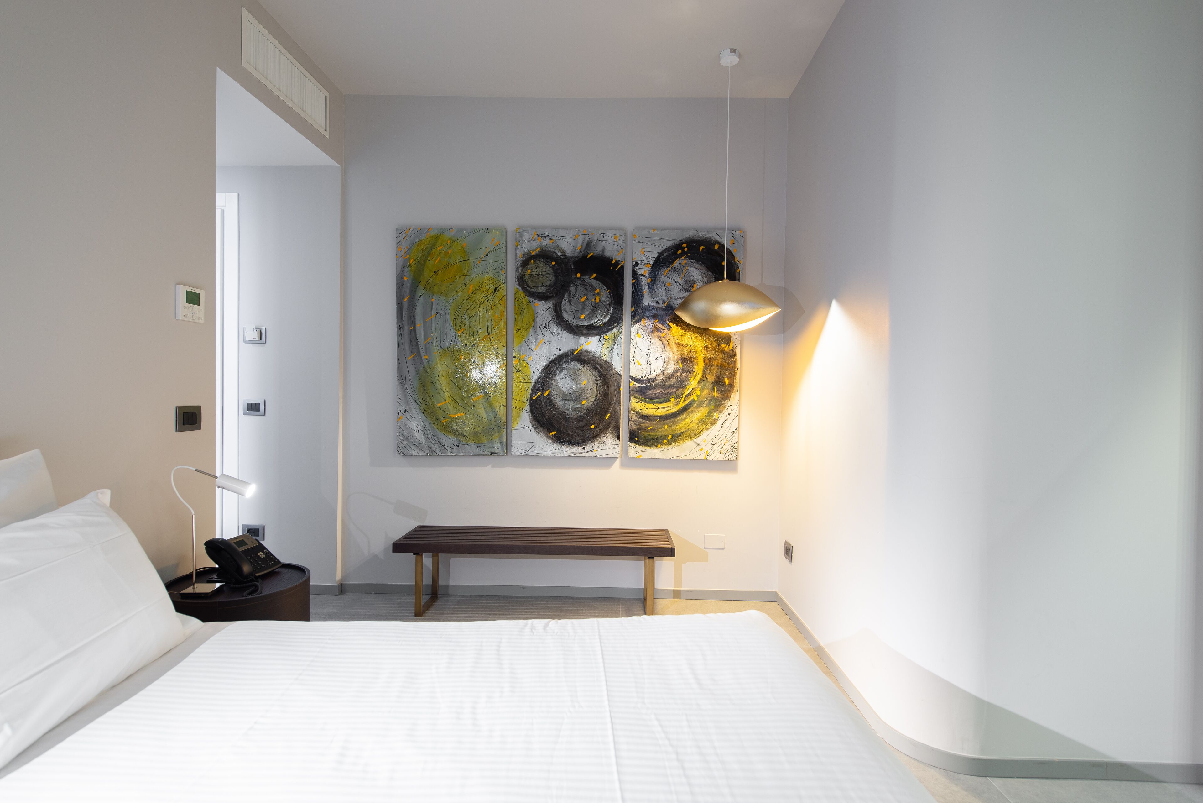 Een hotelkamer met een bed, een bankje, een nachtkastje met een lamp en abstracte muurkunst.