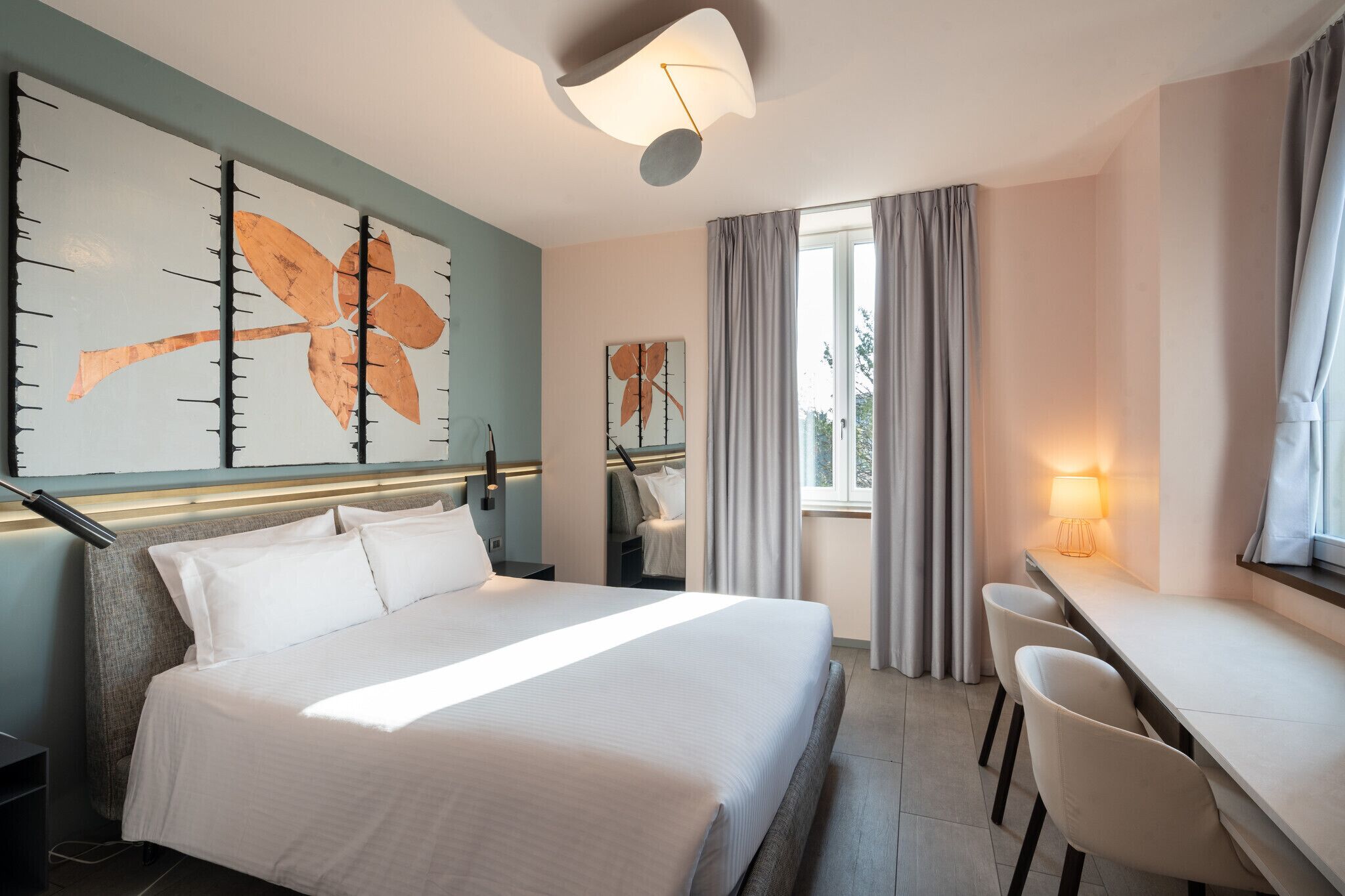 A hotel room with a bed, a desk, and chairs, a large wall art piece, and a window with curtains.