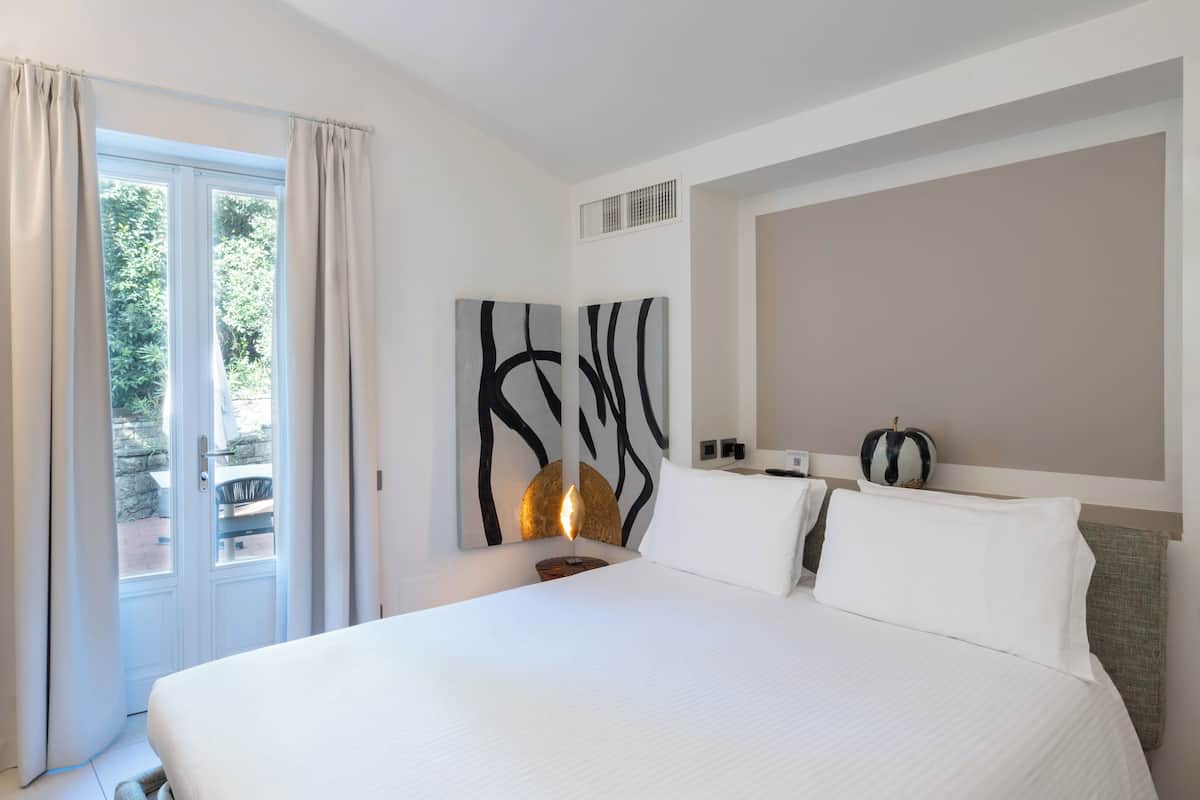 A hotel room with a bed, a large abstract painting, a nightstand, a lamp, and a view of the outdoors through a door.