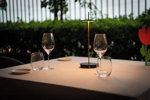 Una mesa con tres copas de vino y una vela, frente a un fondo de follaje y una valla blanca.