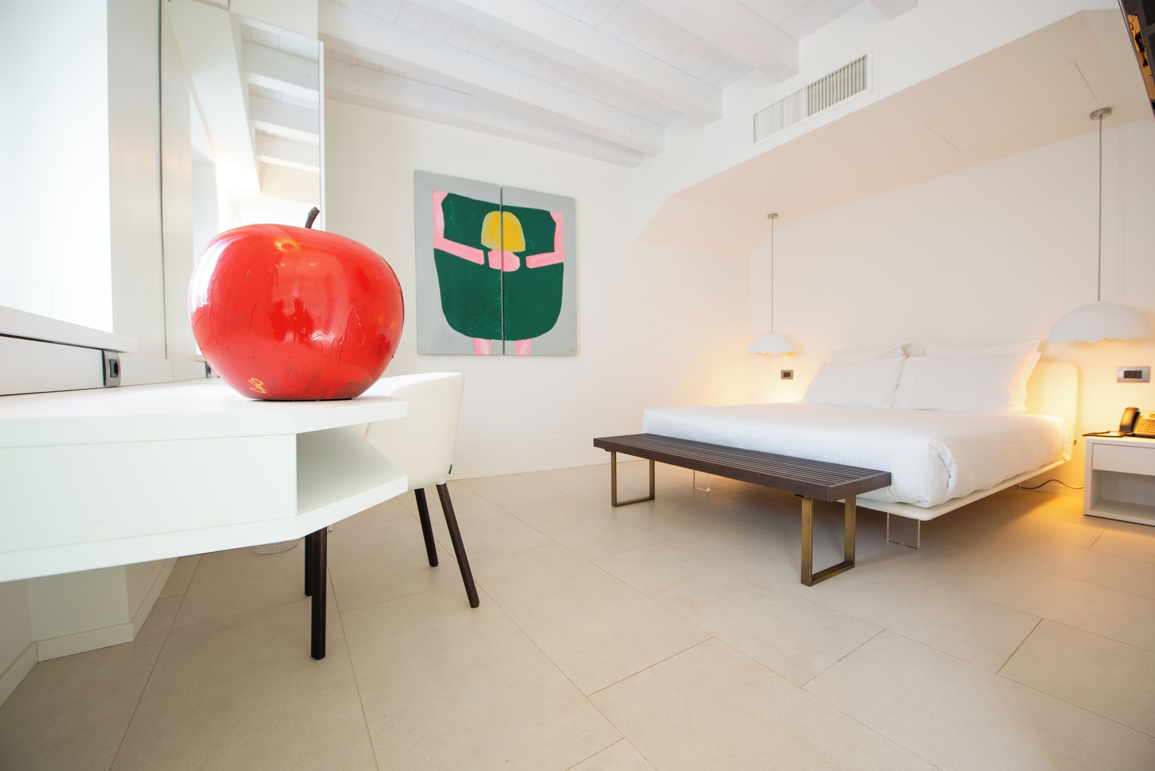 A modern bedroom with a white bed, a desk with a red apple, a bench, and a painting on the wall.