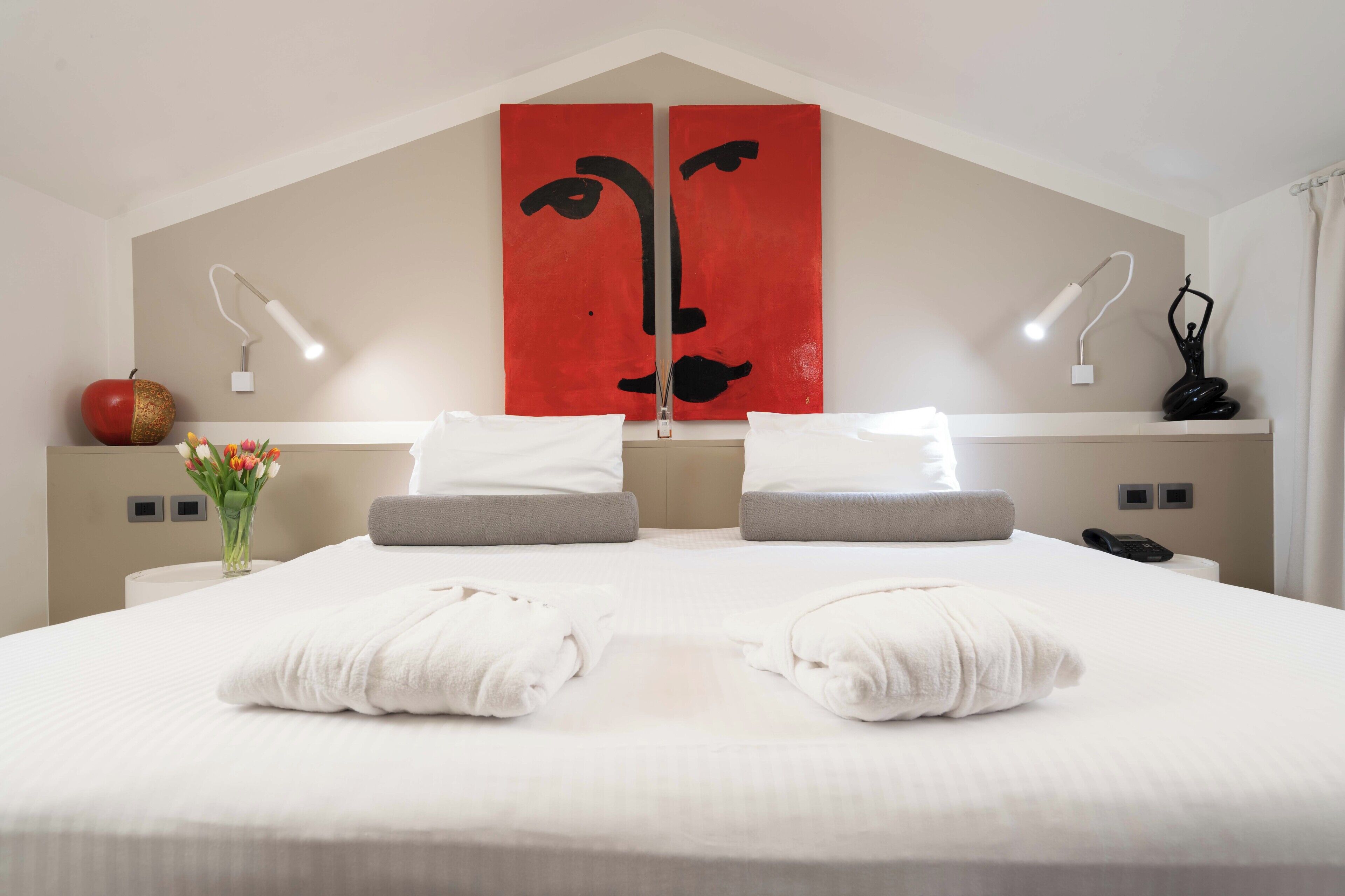 A hotel room with a bed, two bedside lamps, a vase with flowers, a fruit bowl, and a wall art piece.