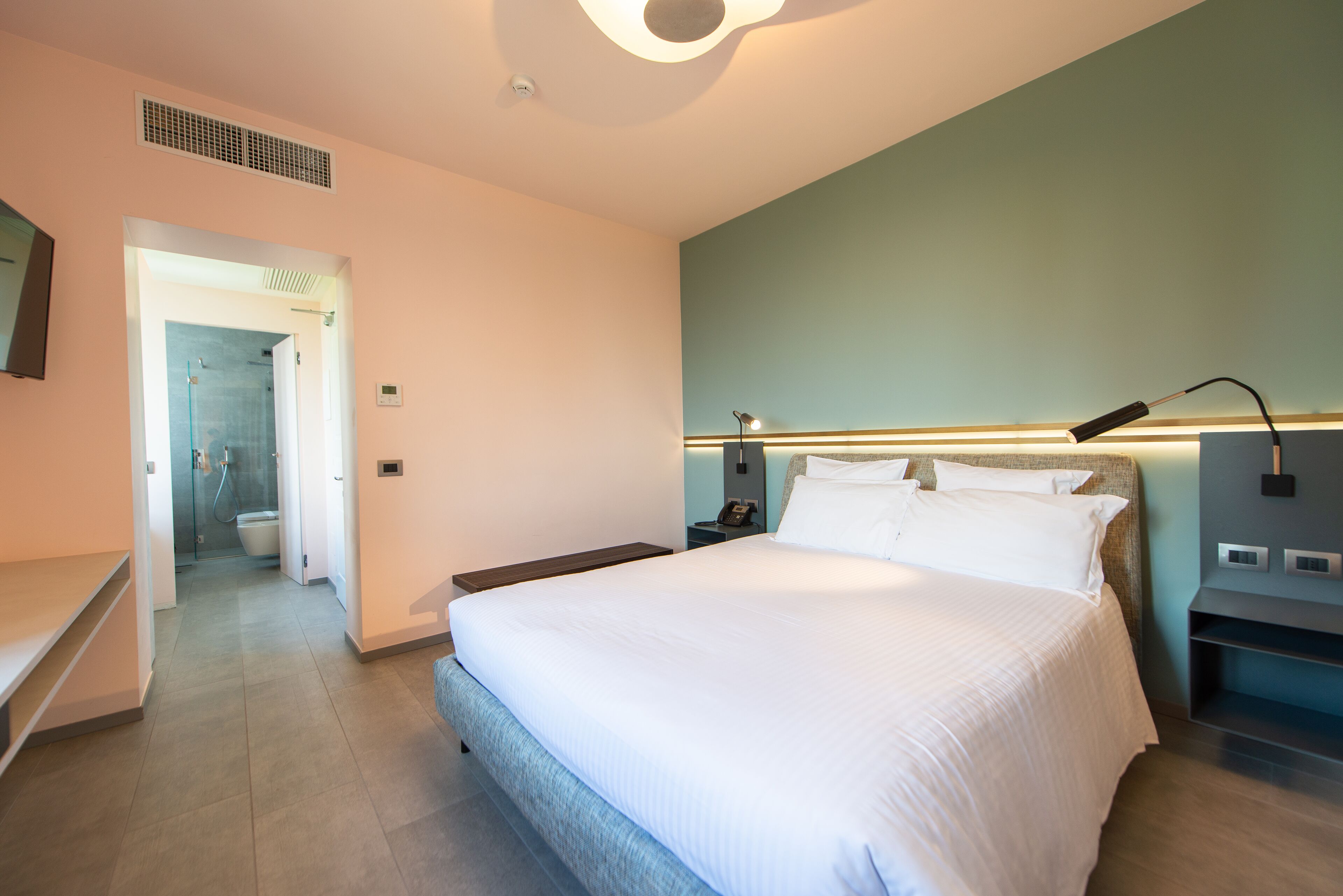 Una habitación de hotel moderna con una cama grande, una mesita de noche, una lámpara de pared y vistas al baño.