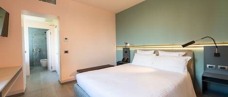 A modern hotel room with a large bed, a bedside table, a nightstand, a wall-mounted lamp, and a view of the bathroom.