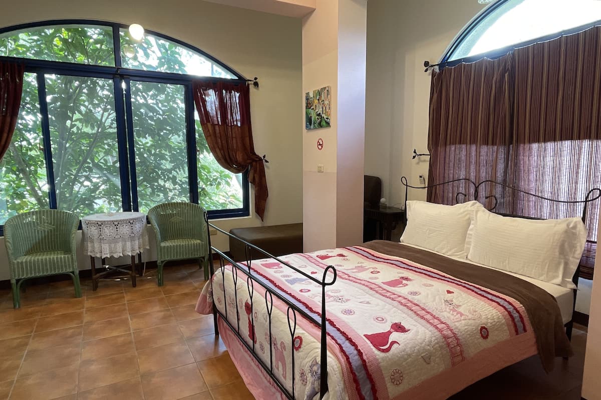 Ein Schlafzimmer mit einem Bett, einem Sessel, einem kleinen Tisch und einem Fenster mit Vorhängen.