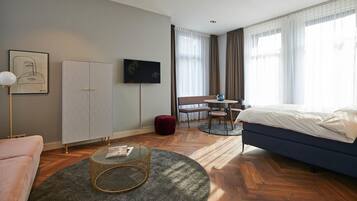 A modern hotel room with a bed, a round coffee table, a sofa, a TV, a wardrobe, and a dining area with a table and chairs.
