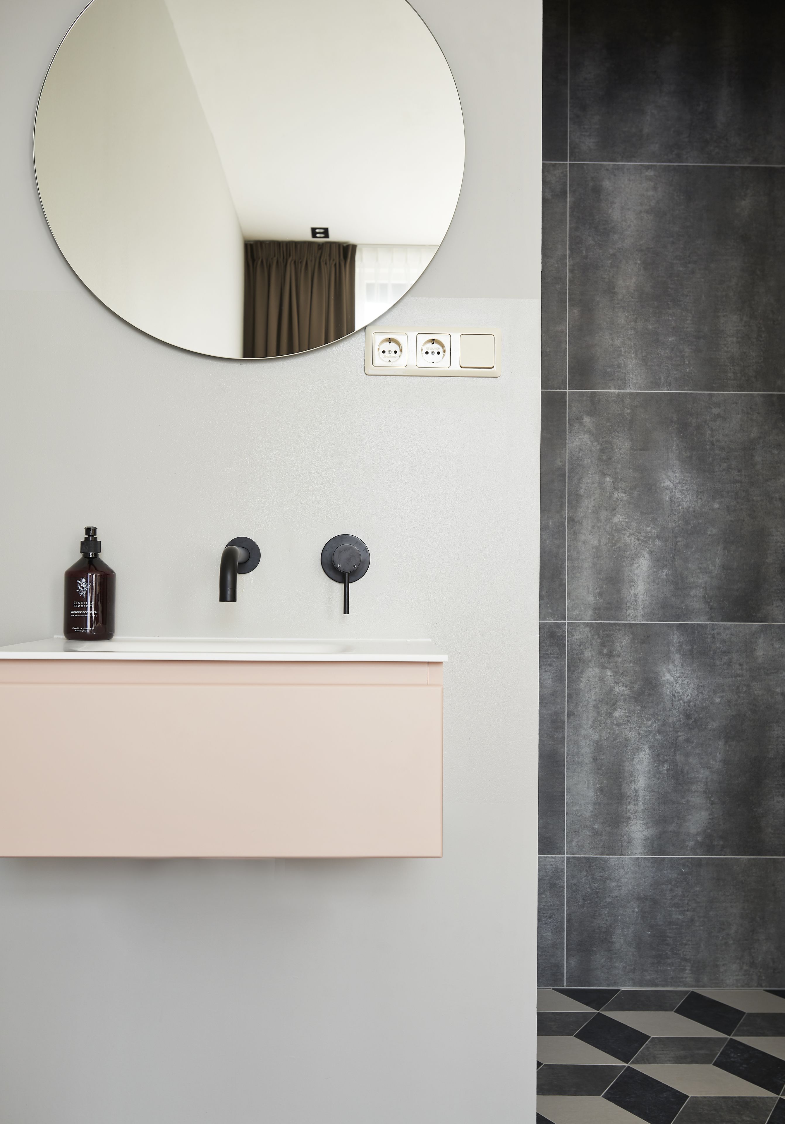 A bathroom with a round mirror, a sink with two faucets, and a bottle of soap.