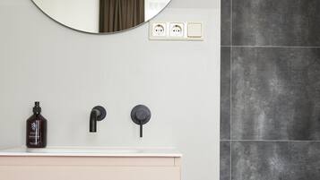 A bathroom with a round mirror, a sink with two faucets, and a bottle of soap.