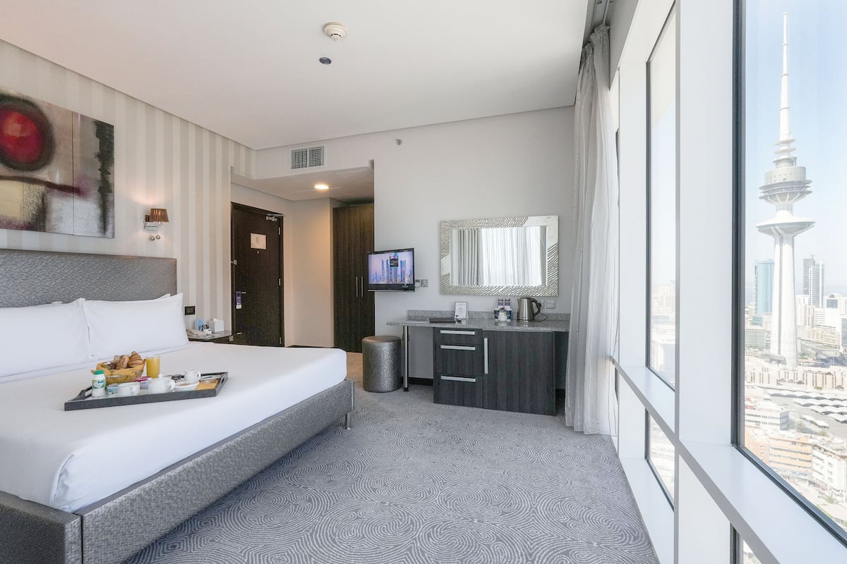 Une chambre d’hôtel moderne dotée d’un grand lit, d’une petite kitchenette, d’une télévision et offrant une vue sur le paysage urbain, notamment sur une tour.