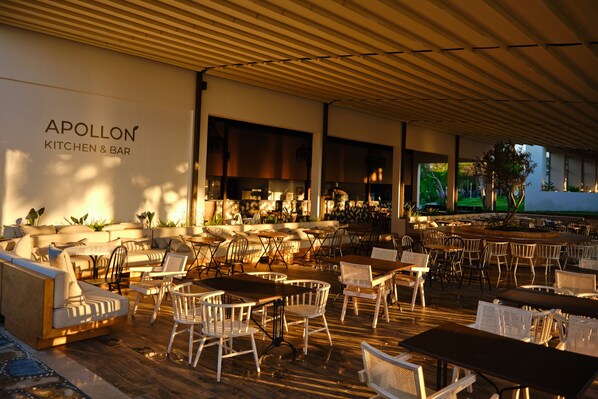 Un área de estar al aire libre con diferentes sillas y mesas, una cocina visible a través de la separación, y un letrero que dice "APOLLON KITCHEN & BAR".