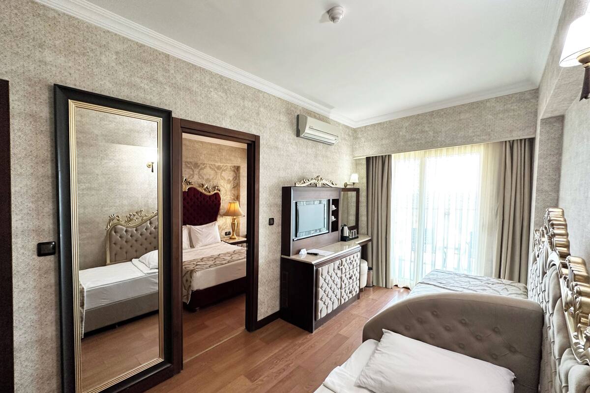 A hotel room with a large mirror, a bed with a tufted headboard, a bedside table with a lamp, a dresser with a mirror, and a television.