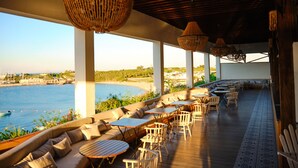 Terraza frente al mar con mesas al aire libre, ventanas amplias y lámparas de mimbre colgantes.