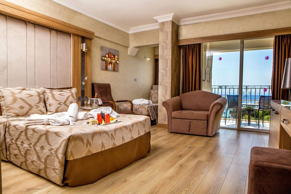 A hotel room with a bed, a chair, a table with a tray of fruit, a balcony with a view, and a painting on the wall.