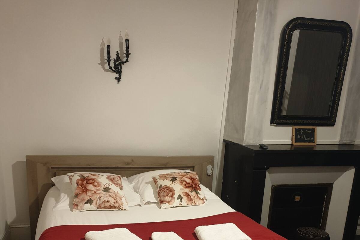 A neatly made bed with a red blanket and three white towels, a wall-mounted candelabra, and a mirror above a fireplace.