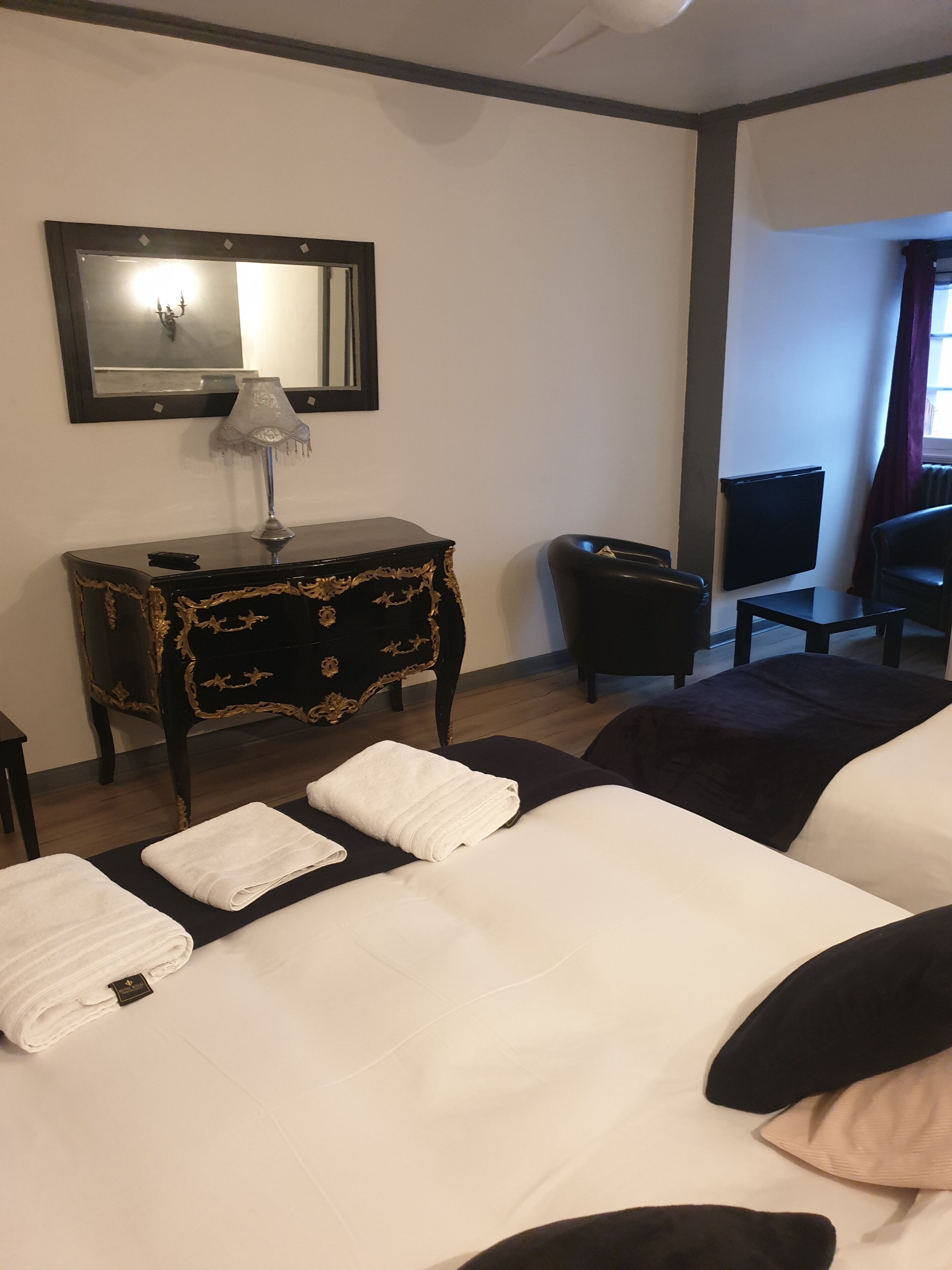 A hotel room with a large bed, a small desk with a lamp, a chair, a radiator, and a window with curtains.