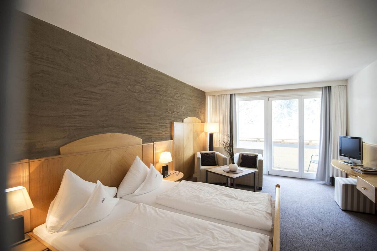 Quarto de hotel com uma cama grande, uma escrivaninha com televisão, uma cadeira e uma janela com cortinas.