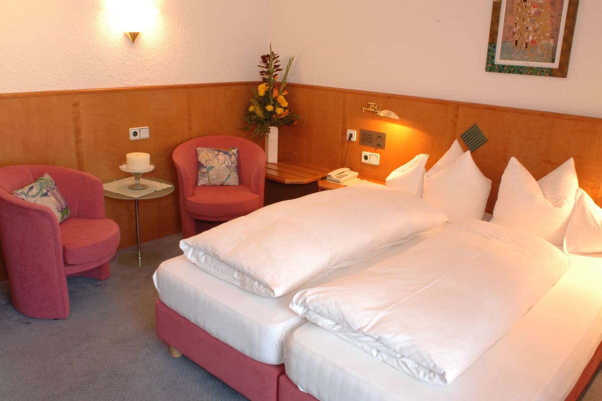 Quarto de hotel com uma cama grande, duas cadeiras, uma mesa pequena com abajur, um vaso com flores e um telefone fixado na parede.