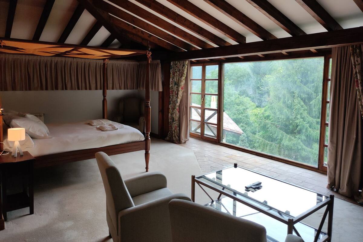 A bedroom with a four-poster bed, a chair, a glass table, and a large window with a view of trees.