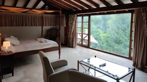A bedroom with a four-poster bed, a chair, a glass table, and a large window with a view of trees. - Viviendas Rurales El Covaju (Cabezon de Liebana)