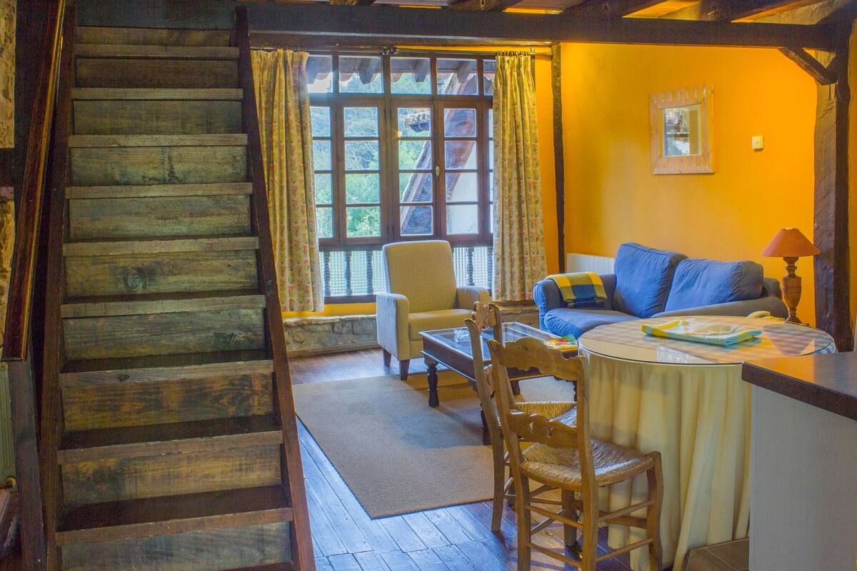 A cozy cabin interior with a wooden staircase, a sofa, a table, and a window with curtains.