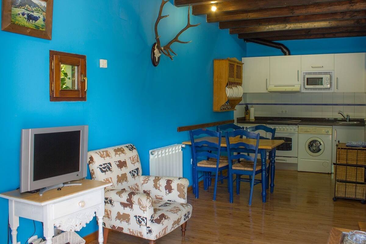 A living room with a blue wall, wooden ceiling, a television, patterned armchair, blue chairs, and a kitchen area with a microwave and washing machine.
