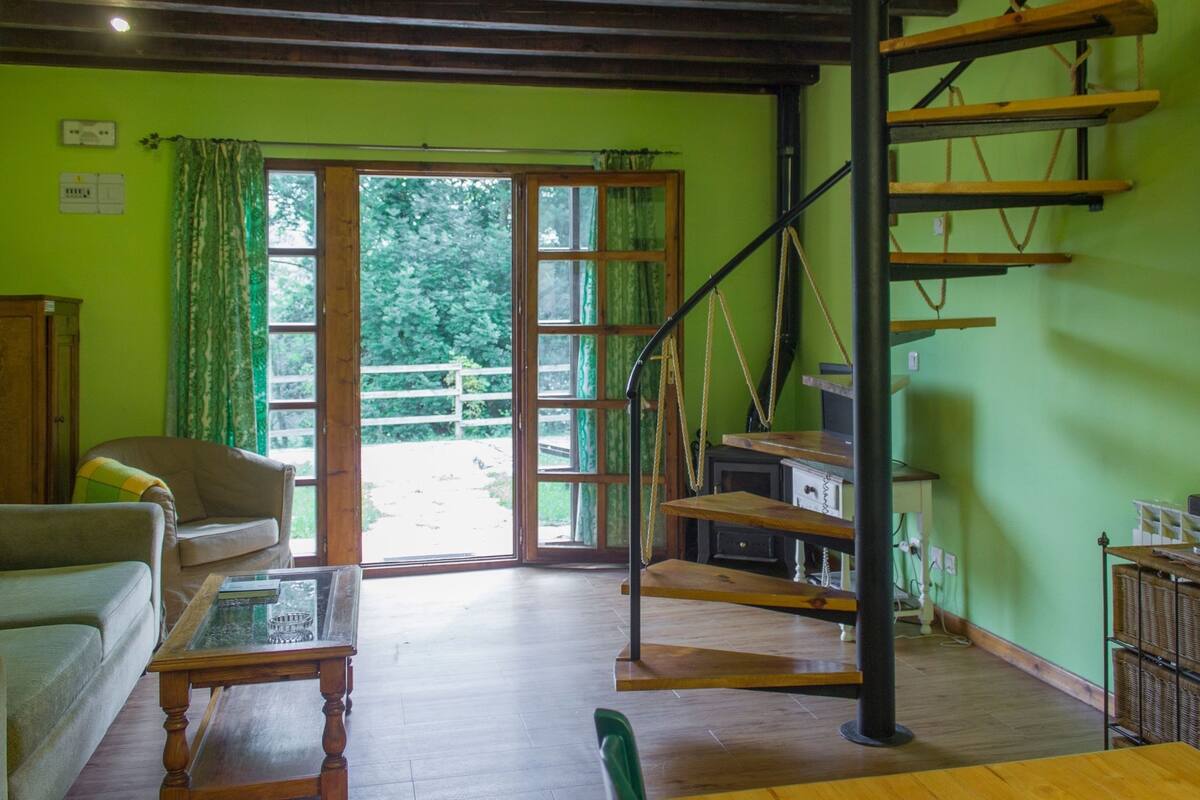 A spacious living area with a wooden staircase, a sofa, a coffee table, and a view through a glass door to an outdoor deck.