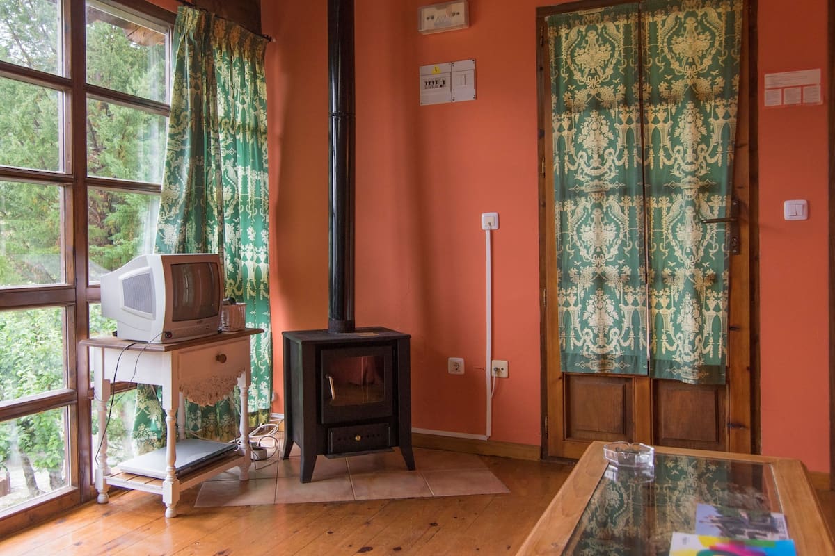 A room with a wood-burning stove, a television on a small table, and a large window with curtains.
