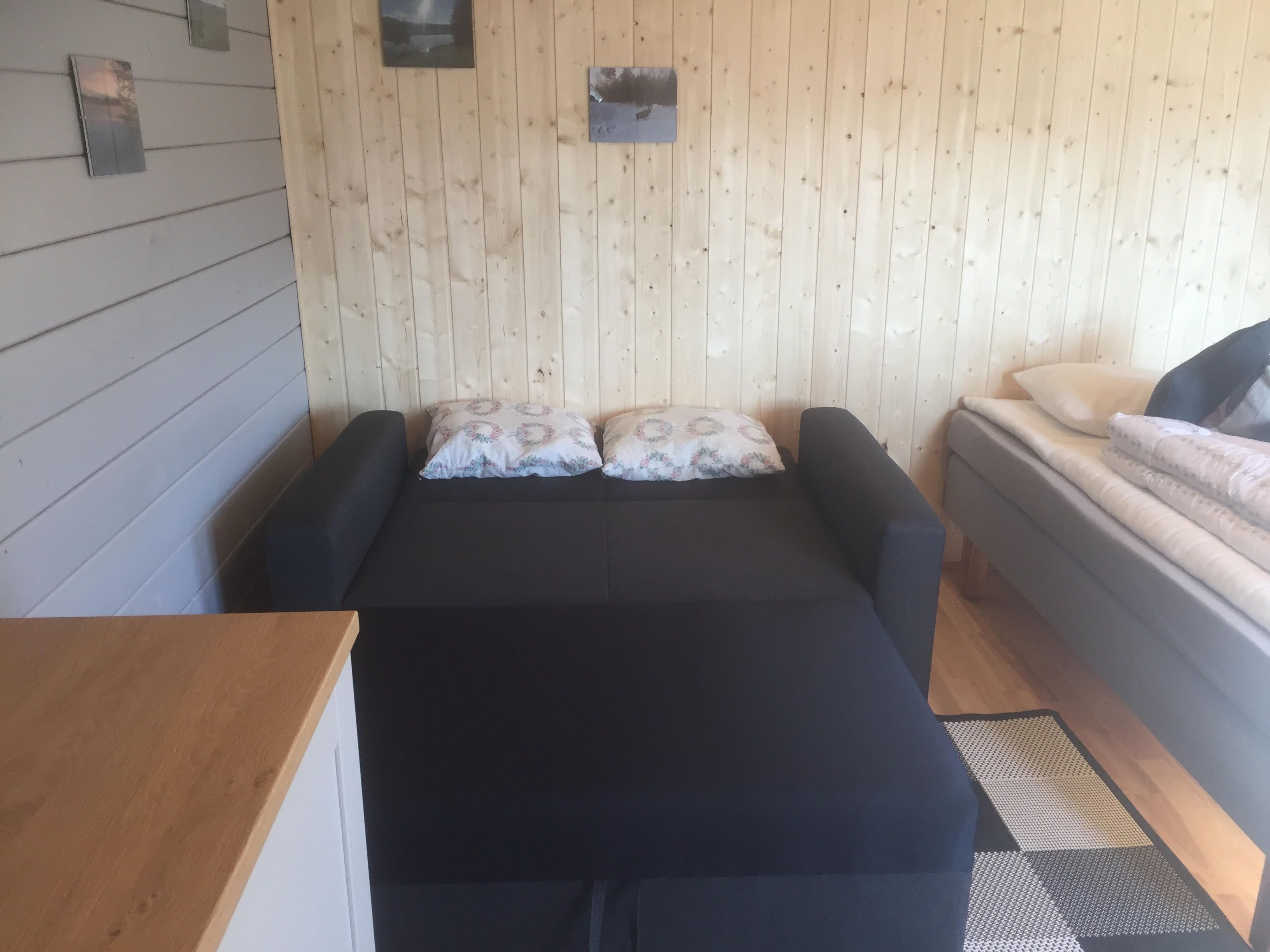 Ein Zimmer mit einer Holzwand, einem Bett mit grau-weißer Matratze, zwei Kissen, einem kleinen Tisch und einem Teppich.