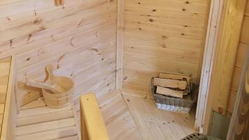 A wooden cabin interior with a sloped ceiling, a wooden bench, a small wooden table, a basket, and a stack of firewood.