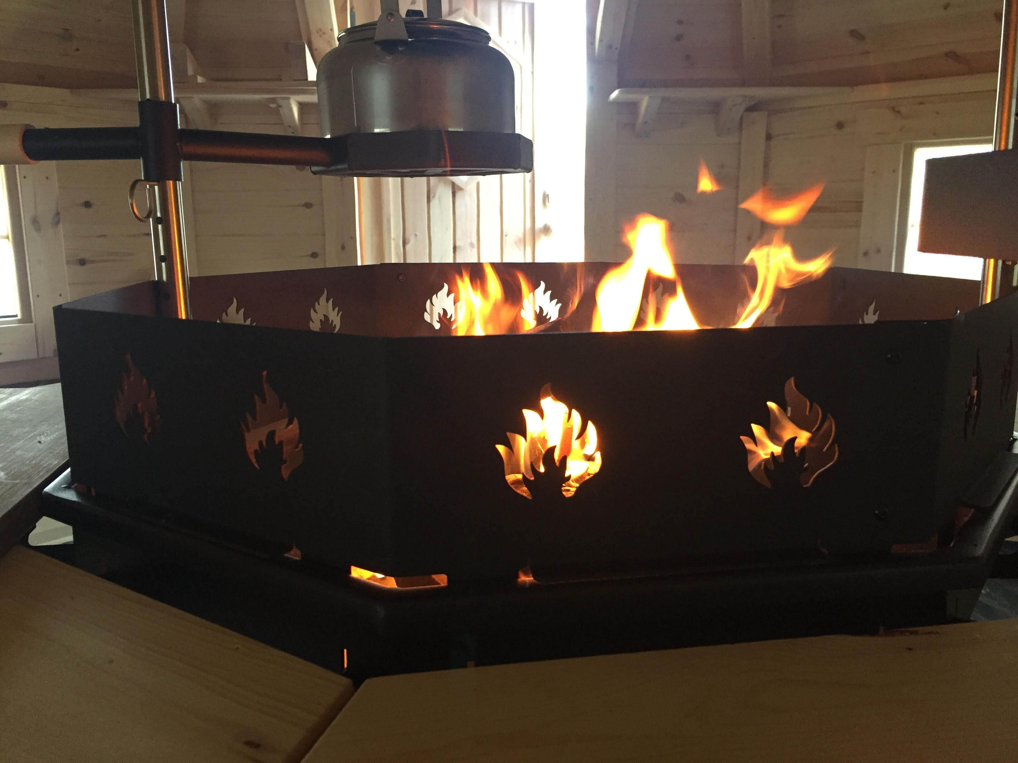 Een metalen vuurkorf met vlammen, geplaatst in een houten kamer met een raam.