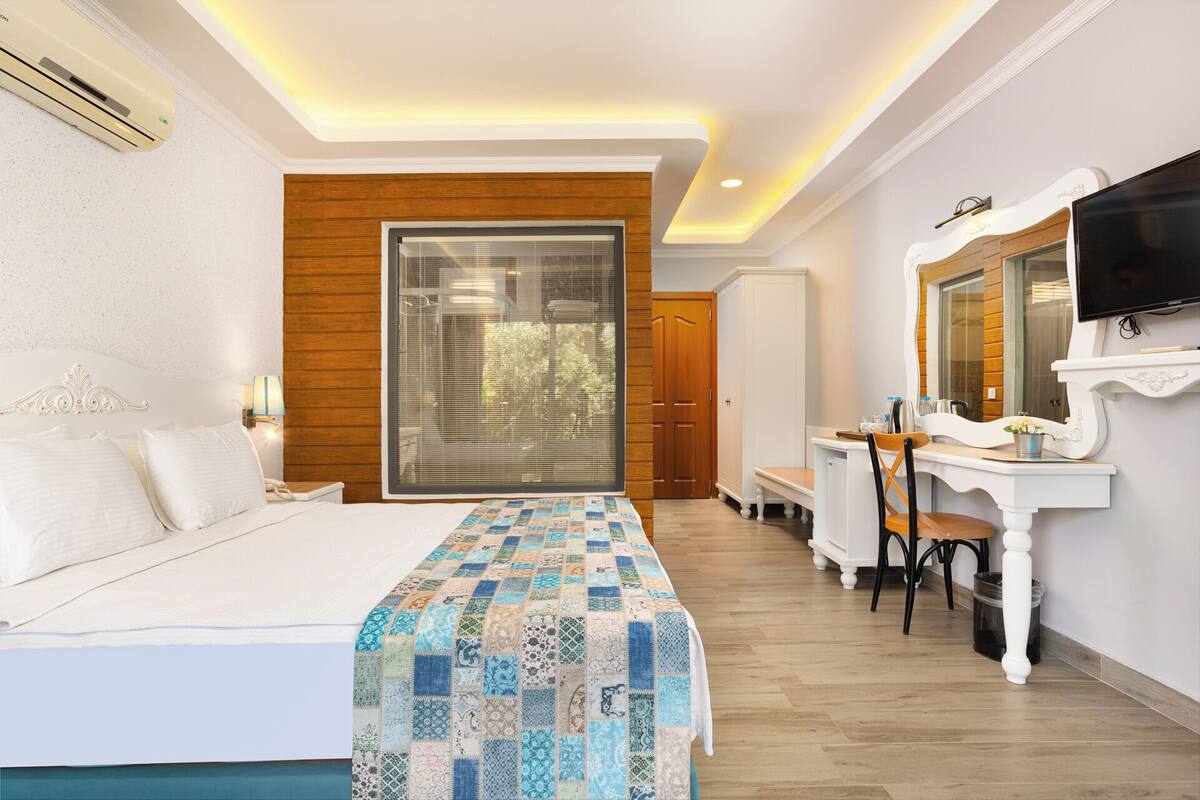 A hotel room with a bed, a desk with a chair, a television, and a large window with blinds.
