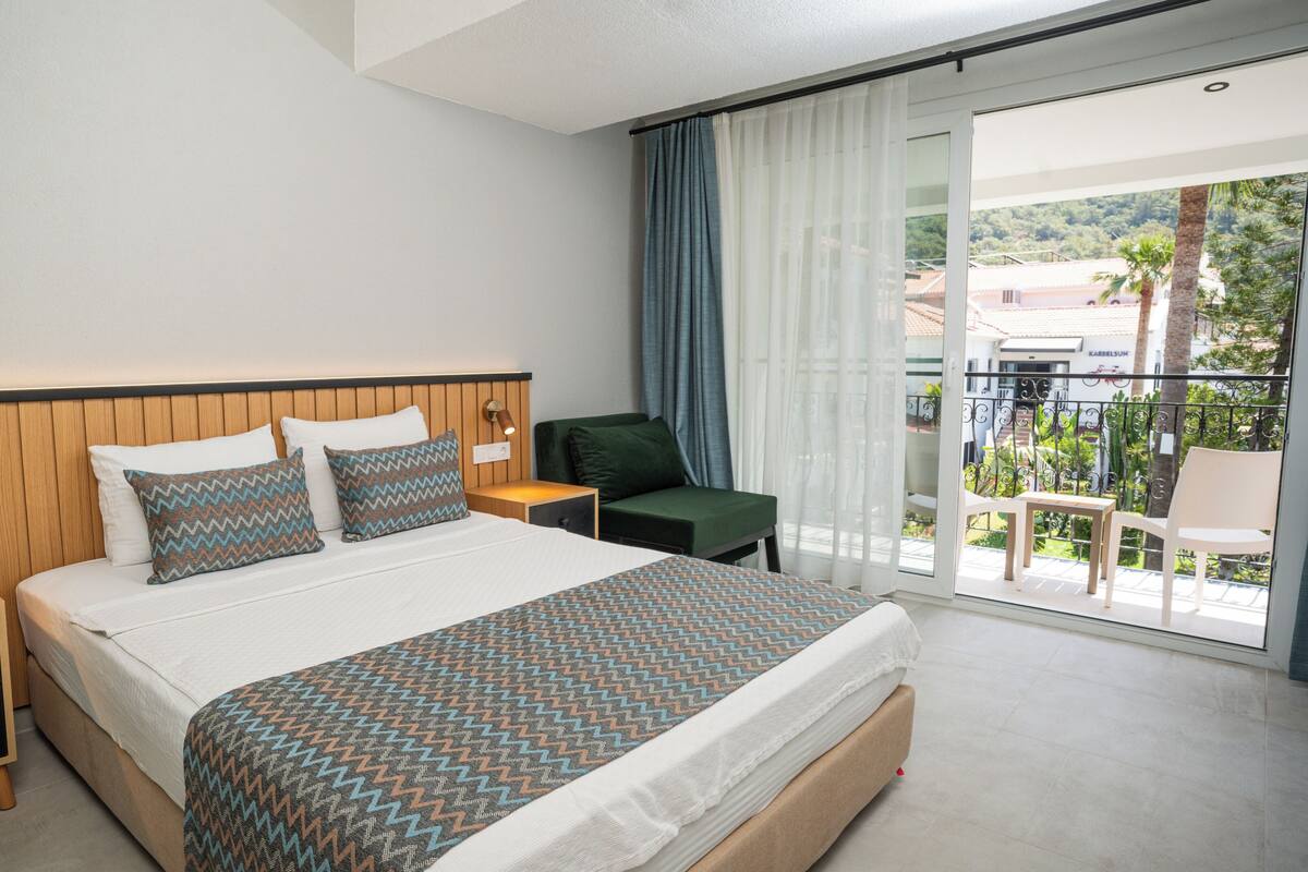 A hotel room with a bed, a nightstand, a chair, and a balcony with a view of the surroundings.