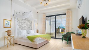 A spacious bedroom with a large bed, a green chair, a wooden desk, and a view through a glass door. - Hotel Karbelsun (Fethiye)
