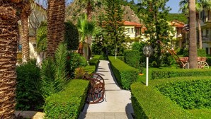 A pathway lined with palm trees and neatly trimmed hedges, leading to a building with a red-tiled roof. - Hotel Karbelsun (Fethiye)