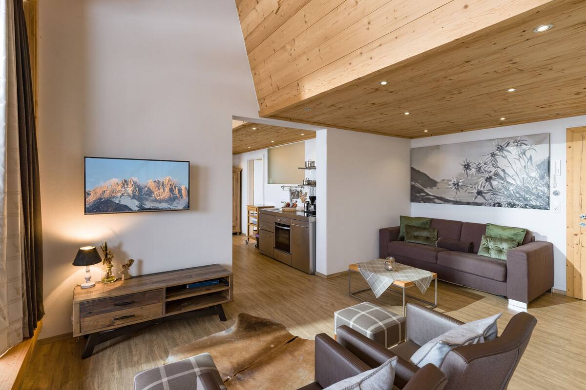Un vaste espace de vie doté d’un plafond en bois, d’un ensemble de canapés, d’une table basse et d’un téléviseur fixé au mur.